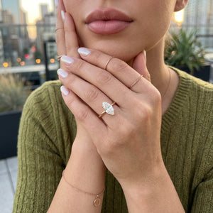 A close-up lifestyle shot of a woman wearing a rose gold ring with a marquise-cut clear gemstone on her finger, posing with her hands near her face. She is wearing an olive-green knit sweater, and a blurred city skyline is visible in the background - J.Collins Jewelry