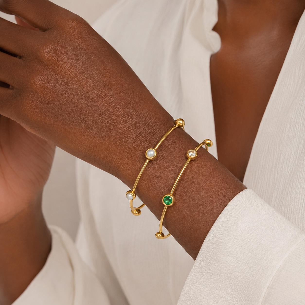 Gold bangles with pearl and emerald details on a model's wrist wearing a white sleeve.