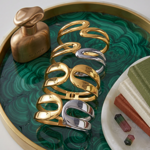 Gold and silver Omega Flow Bracelets on a green tray with a bronze perfume bottle and books nearby.