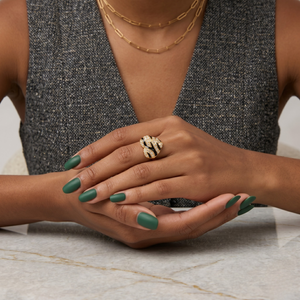 Gold snake-like ring on woman's hand with green nails, worn with a gray textured vest and layered necklaces.