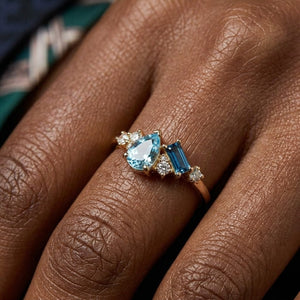 Gold ring with pear and baguette-cut blue stones, accented by small white gems on a hand.