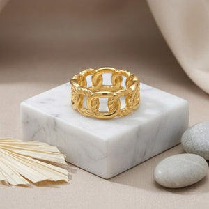 Gold anchor cage ring on a marble stand with pebbles and soft fabric background