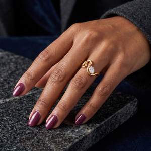 Gold gemstone ring on a person's hand with plum colored nails, displayed on a dark marble surface
