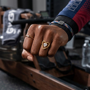 Gold heart-shaped Couture Amour Ring on hand, gym setting, wristband visible, emphasizing active lifestyle.