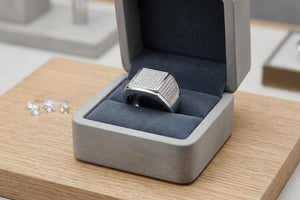 Silver Alpha Crest Ring with a square, diamond-encrusted top in a gray jewelry box on a wooden surface.