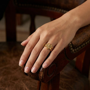 Gold wildflower petal ring on a woman's hand resting on a wooden chair arm.