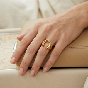 Gold horseshoe ring with diamond accents worn on a hand resting on beige leather car interior.