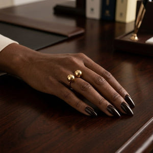 Model wearing a statement gold open bypass ring with two polished spheres resting on a dark wooden desk | J.Collins Jewelry