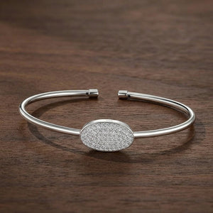 A polished silver cuff bracelet with a central oval pavé-set stone cluster, displayed on a dark brown wooden surface.
