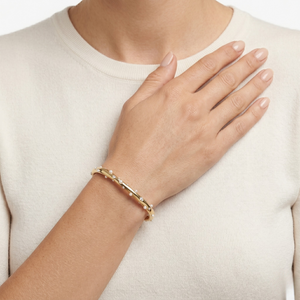 Gold cuff bracelet with bead accents on person's wrist, hand on chest, cream clothing background