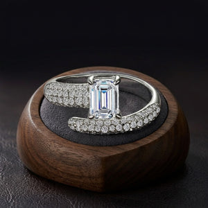 An emerald-cut diamond ring with pavé-set diamonds on a silver-toned band, displayed on a dark textured ring stand inside a round wooden box against a dark background - J.Collins Jewelry