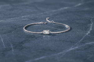 A white gold tennis bracelet with a rectangular baguette-cut center stone, displayed on a dark slate surface - J.Collins Jewelry