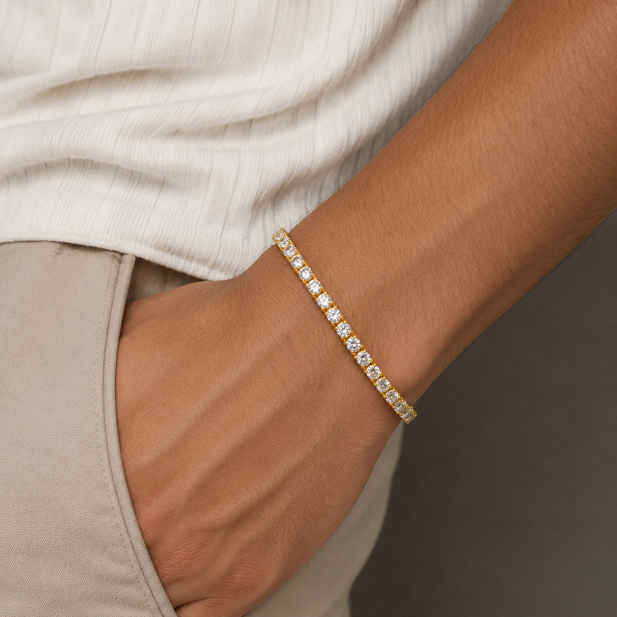 A close-up of a person's wrist wearing a delicate gold tennis bracelet with small clear stones, styled with a cream ribbed top and beige trousers - J.Collins Jewelry