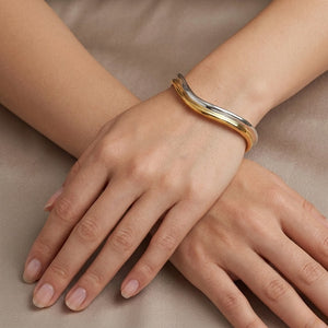 Bi-Color Arc Bracelet in gold and silver on model's wrist with hands elegantly crossed on beige fabric surface.