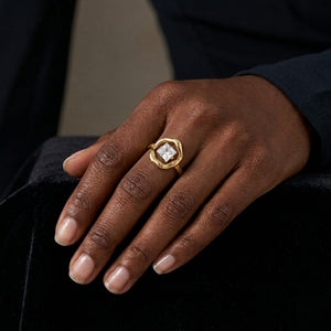 Gold ring with hexagonal design on hand, featuring a large square gemstone; black fabric backdrop.