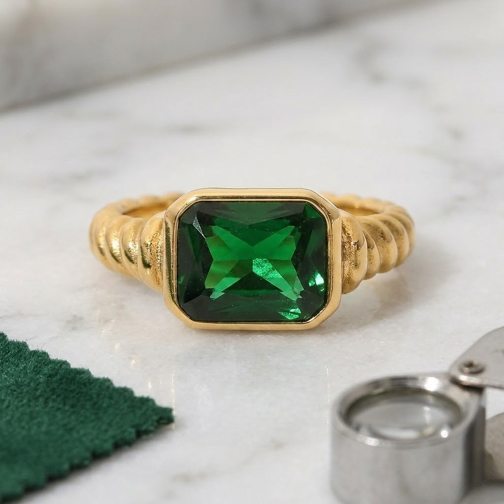 Gold ring with square-cut green gemstone and ribbed band style on marble surface with green cloth and small magnifier.