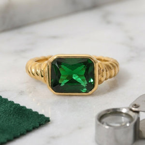 Gold ring with square-cut green gemstone and ribbed band style on marble surface with green cloth and small magnifier.
