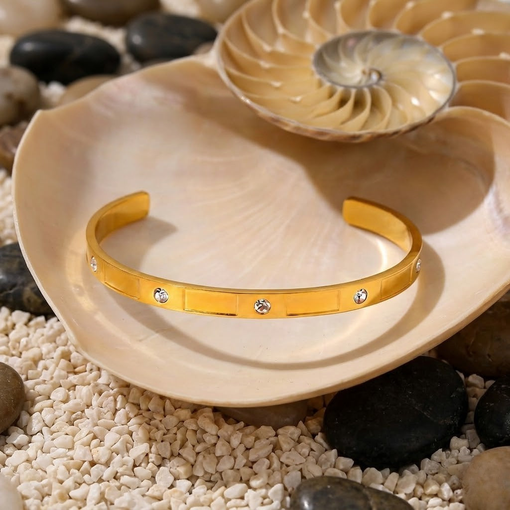 Golden cuff with crystal accents displayed on a seashell surrounded by pebbles on white sand.