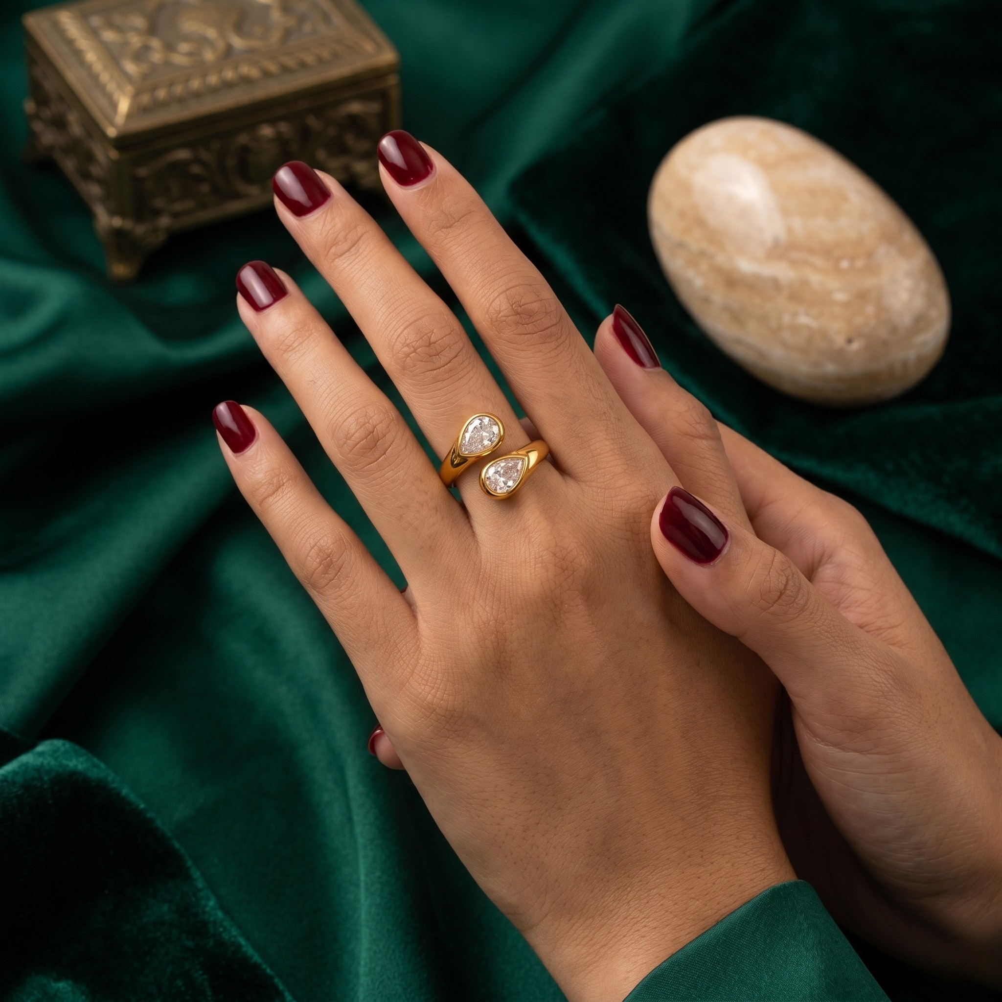 Gold teardrop ring with clear stones on hand with dark red nails, set against green velvet and decorative items