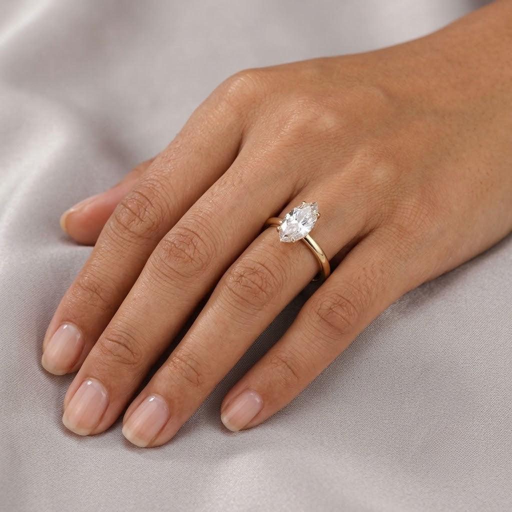 A close-up of a hand wearing a gold ring with a marquise-cut clear gemstone, resting on a soft light gray fabric background - J.Collins Jewelry