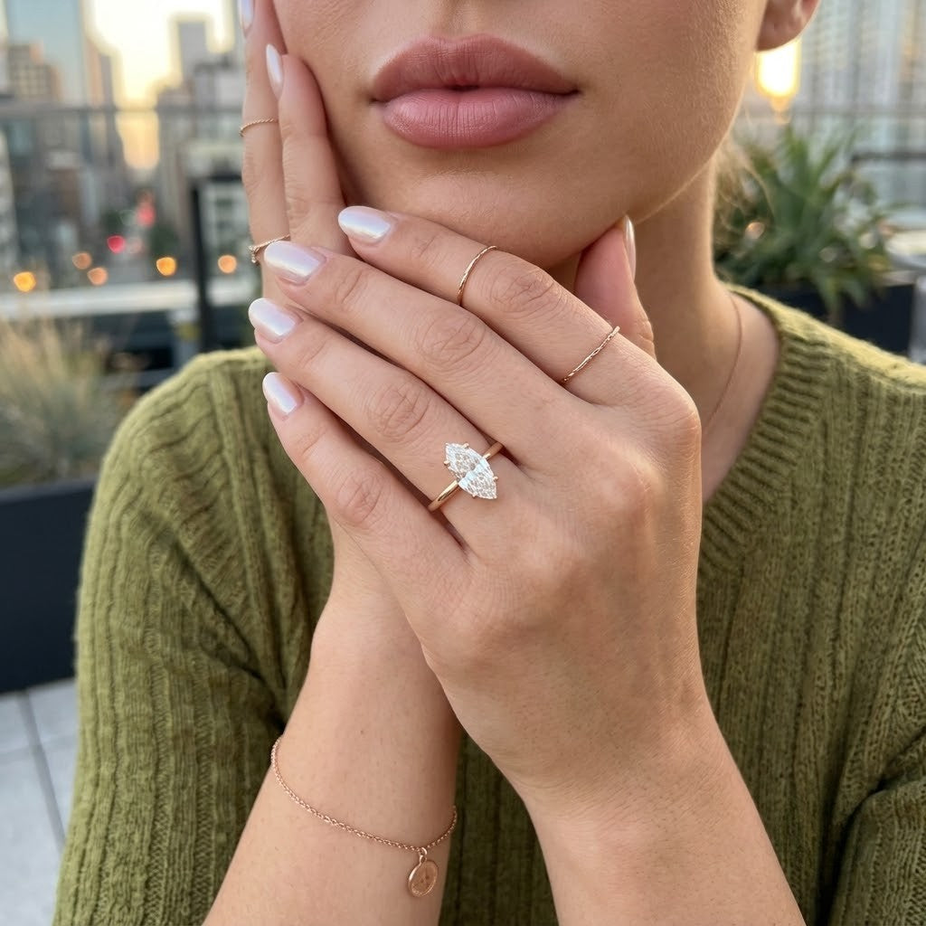 A close-up lifestyle shot of a woman wearing a rose gold ring with a marquise-cut clear gemstone on her finger, posing with her hands near her face. She is wearing an olive-green knit sweater, and a blurred city skyline is visible in the background - J.Collins Jewelry