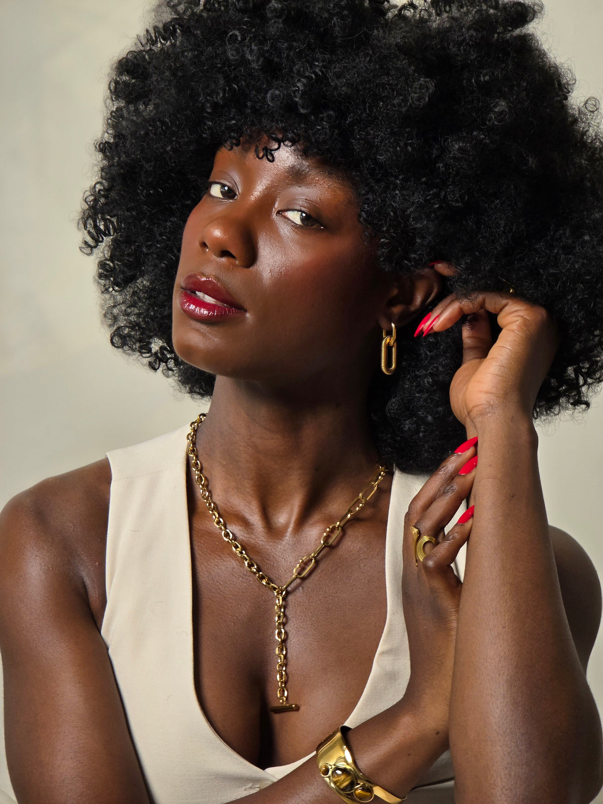A woman with a large black afro hairstyle wearing a cream-colored sleeveless top, gold jewelry, and red nail polish, posing with her hands near her face against a neutral indoor background - J.Collins Jewelry