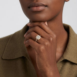 Gold ring featuring a cushion-cut cubic Moissanite center stone surrounded by a halo of round accents worn on finger by model  wearing an olive green collared shirt by J.Collins
