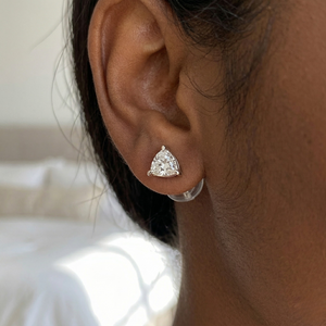 A close-up photograph of a person with dark skin wearing a large, sparkling trillion-cut diamond stud earring on their left earlobe. The earring is set in a simple white gold or platinum bezel setting and secured with a clear plastic butterfly backing, visible from the back. The setting has two tiny details at the bottom of the triangle point. The person has dark hair that is pulled back, showing the details of their ear. The background is a gently blurred indoor space with white bedding, out of focus.