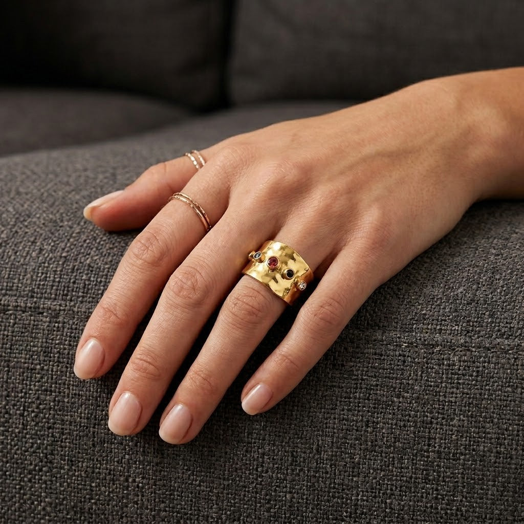 Gold Aura Gemstone Band with colorful stones on a hand, paired with delicate rings, on a gray couch background