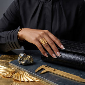 18K gold wing loop ring on hand holding black clutch, paired with gold bracelet and surrounded by decorative elements