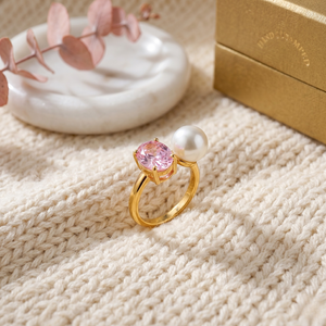 Gold ring with pink gemstone and pearl on a textured fabric background, styled with a leaf accent and gold box.