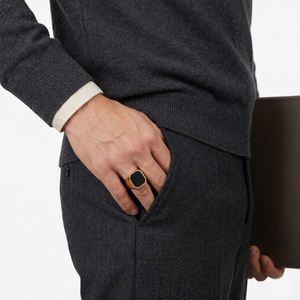 Man wearing gold onyx signet ring with hand in pocket, holding brown portfolio, wearing dark sweater and trousers.