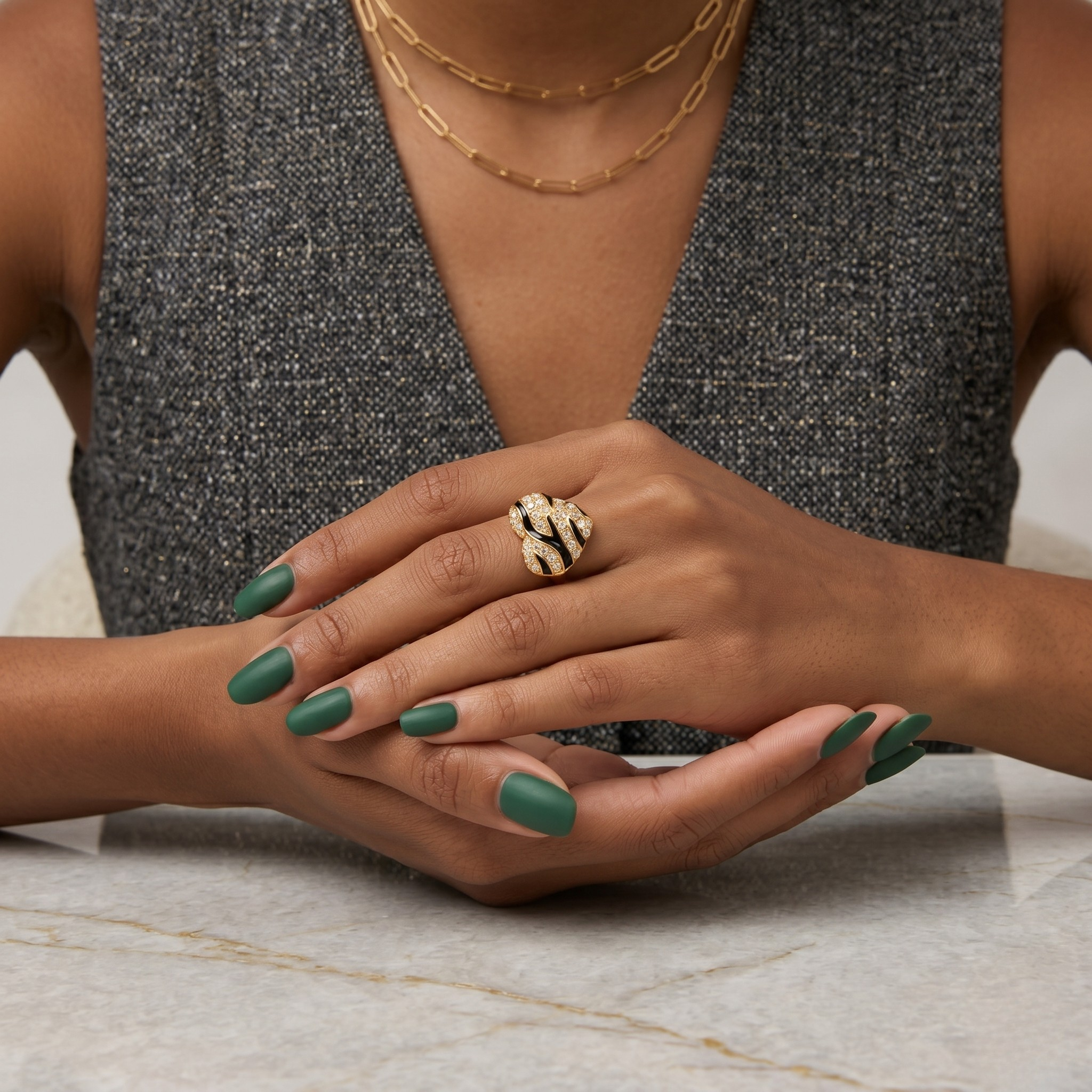 Gold snake-like ring on woman's hand with green nails, worn with a gray textured vest and layered necklaces.