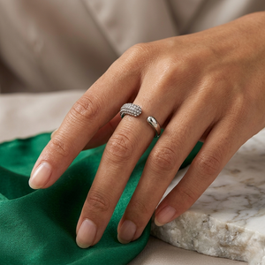 Hand wearing silver curved pavé ring with diamonds, resting on marble, green fabric nearby