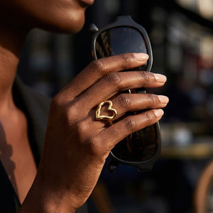 Gold hollow heart ring on hand holding black sunglasses, outdoor lifestyle setting