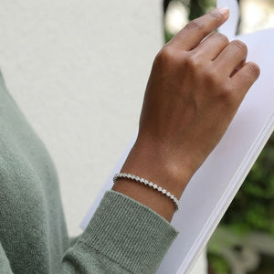Classic single-row sterling silver tennis bracelet set with round cubic Moissanite crystals worn on wrist by model wearing a sage green sweater holding a book by J.Collins
