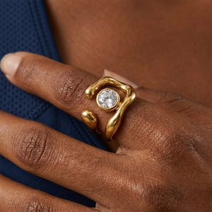Gold kinetic ring with a round crystal on a hand, featuring a fluid, organic design and worn by a person.