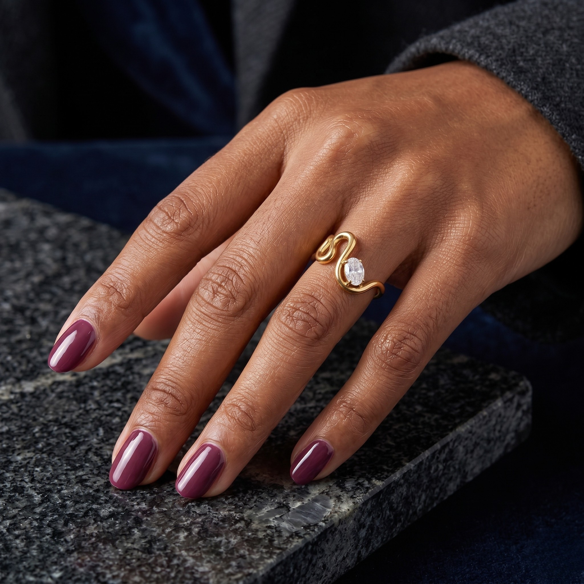 Gold gemstone ring on a person's hand with plum colored nails, displayed on a dark marble surface