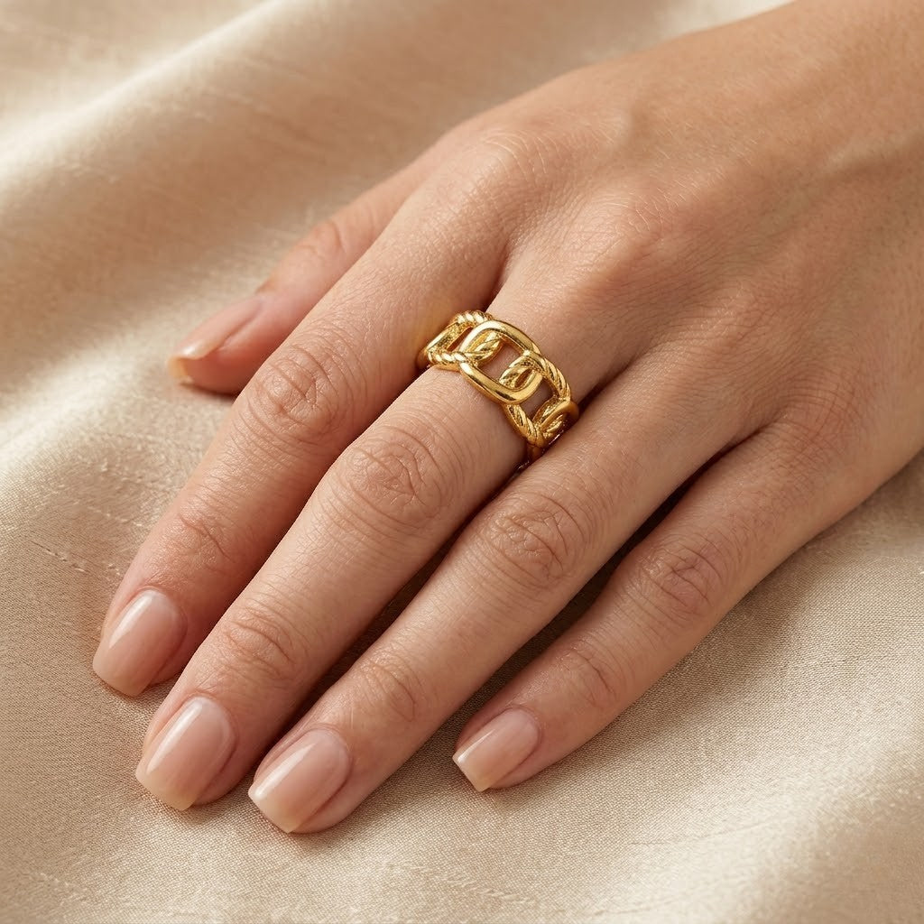 Gold anchor cage ring on hand with neutral nails on beige satin fabric