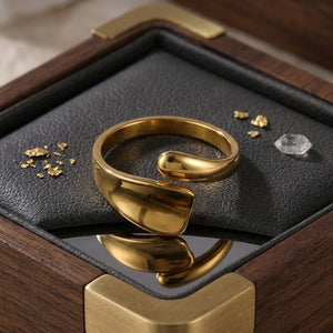 Gold wrap-around ring with a unique pivot design displayed on a leather box with gold flakes and a clear gem nearby.