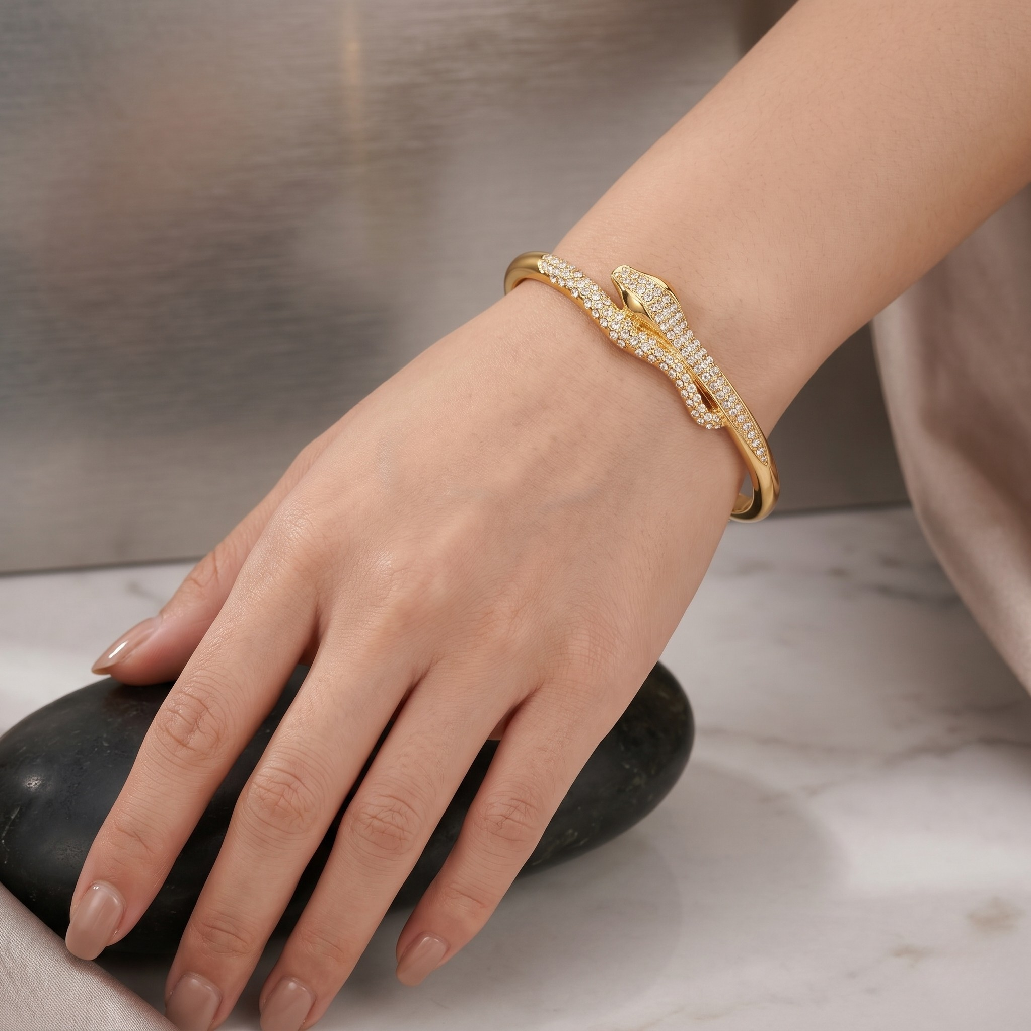 Gold sculptural snake open cuff adorned with crystals on a woman's wrist, hand resting on a dark stone.