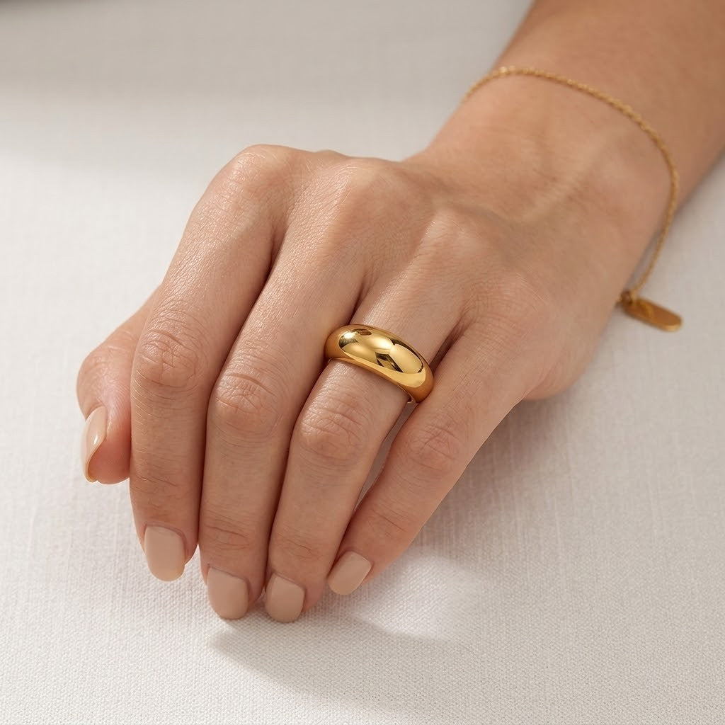 Gold dome-style ring on hand with neutral nail polish, delicate bracelet on wrist, soft fabric background.