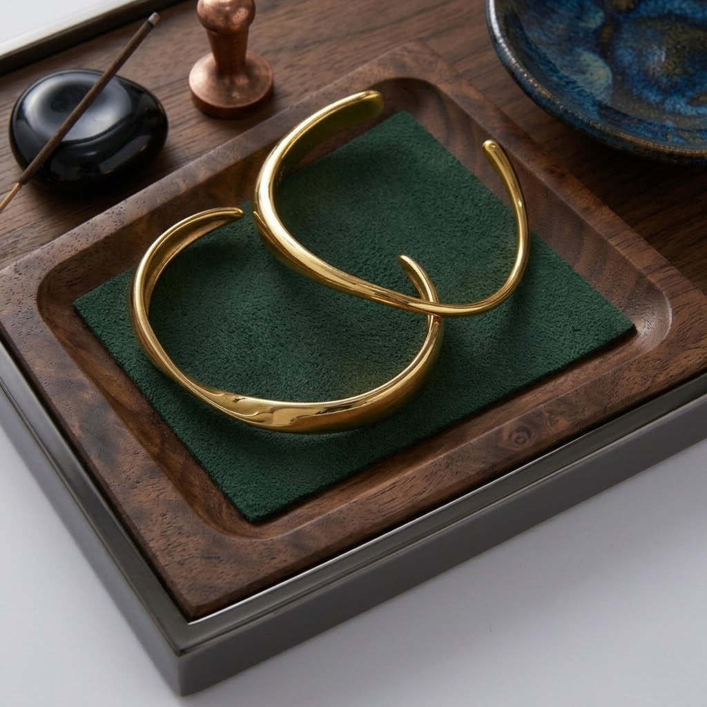 Gold infinity curve bangles on a wooden tray with green fabric, featuring a dark ceramic dish and decorative items.