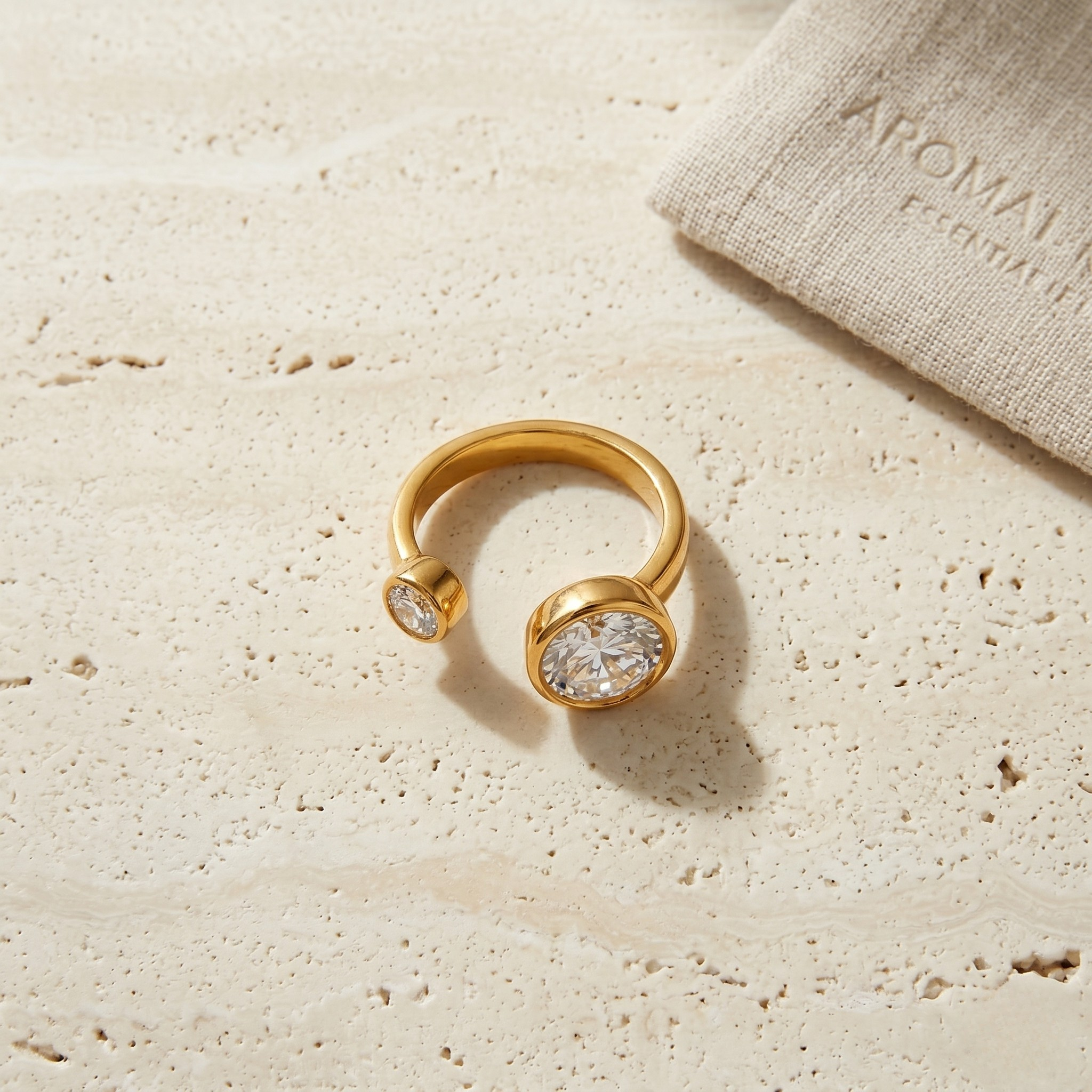 Gold open steel ring with two diamonds on a textured stone surface, near a linen pouch