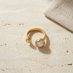 Gold open steel ring with two diamonds on a textured stone surface, near a linen pouch