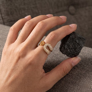 Hand wearing gold and diamond-encrusted horseshoe ring, holding a black stone, on a cushioned surface.