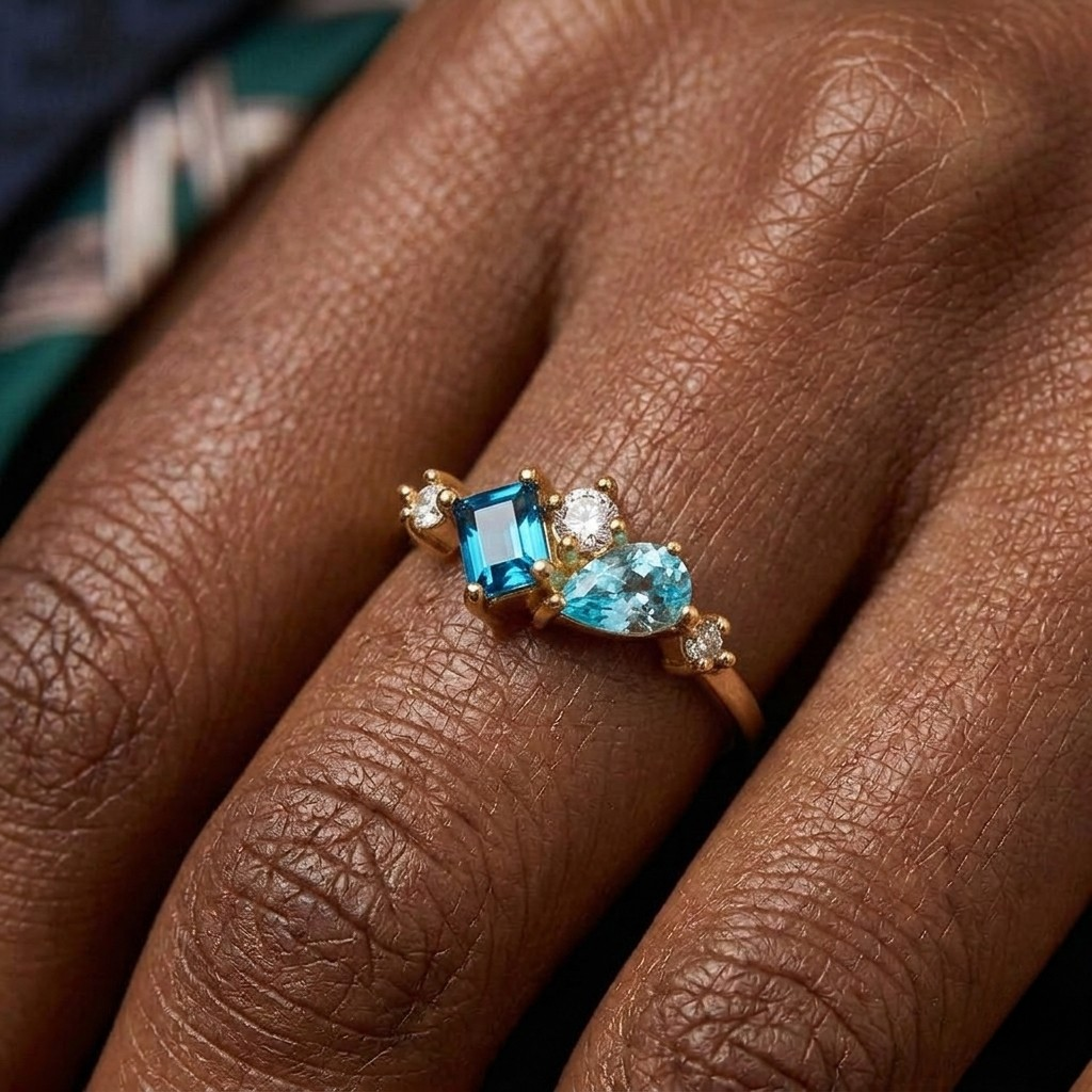 A close-up macro shot of a delicate gold ring worn on a hand. The asymmetric cluster ring features a deep blue emerald-cut gemstone alongside an angled light blue pear-cut gemstone, accented with smaller round clear stones set in gold prongs along the band.
