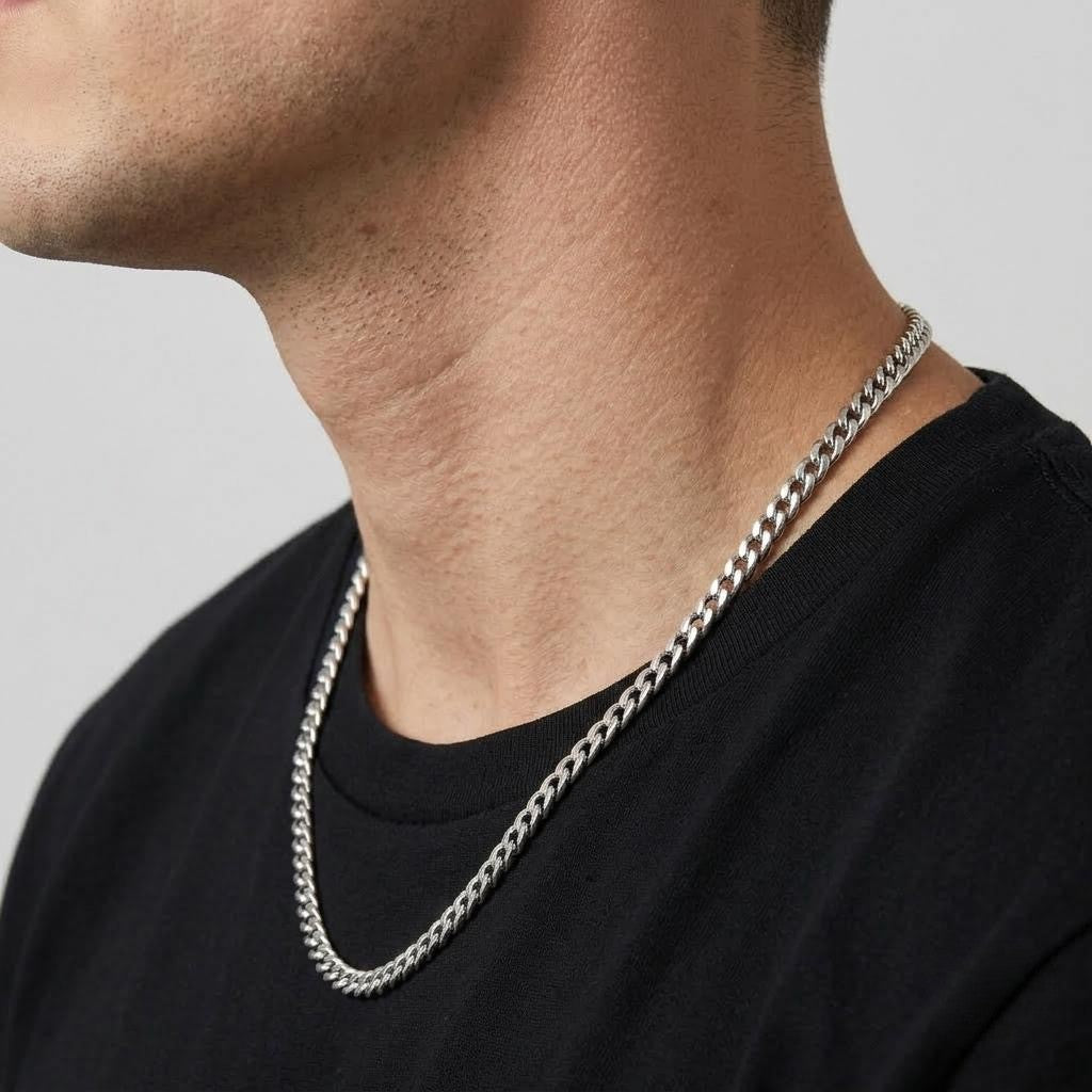A close-up of a man's neck and lower face wearing a silver-toned curb chain necklace over a black t-shirt, set against a plain light gray background - J.Collins Jewelry