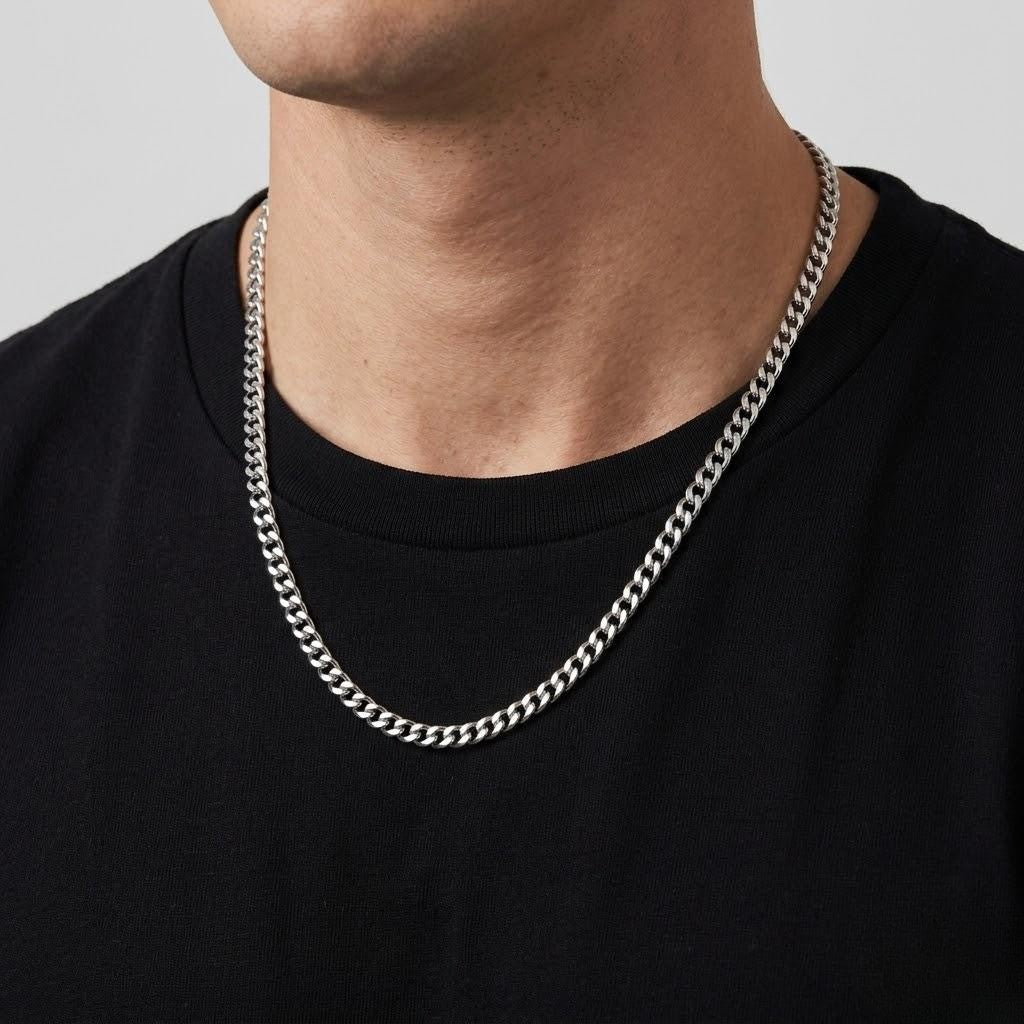 A close-up of a man's neck and chest wearing a silver-toned curb chain necklace over a black t-shirt, against a light gray background - J.Collins Jewelry