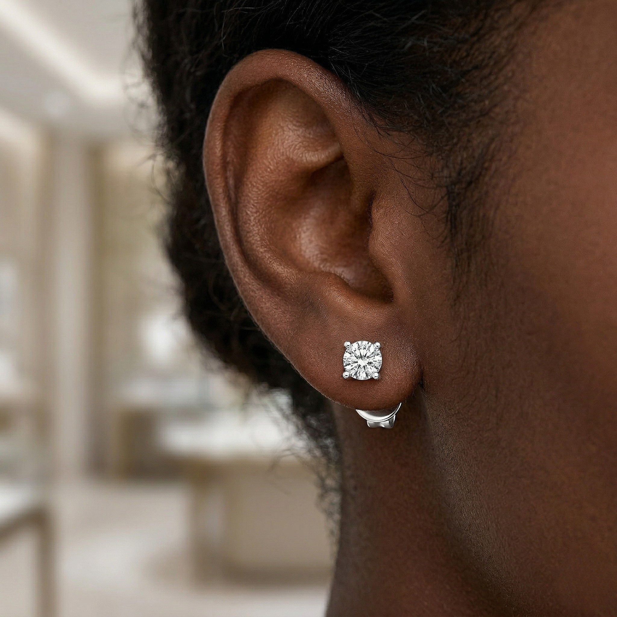 Close-up of a person wearing a diamond stud earring with a blurred indoor background - Diamond Earrings Basket 0.144ct - J Collins Jewelry 
